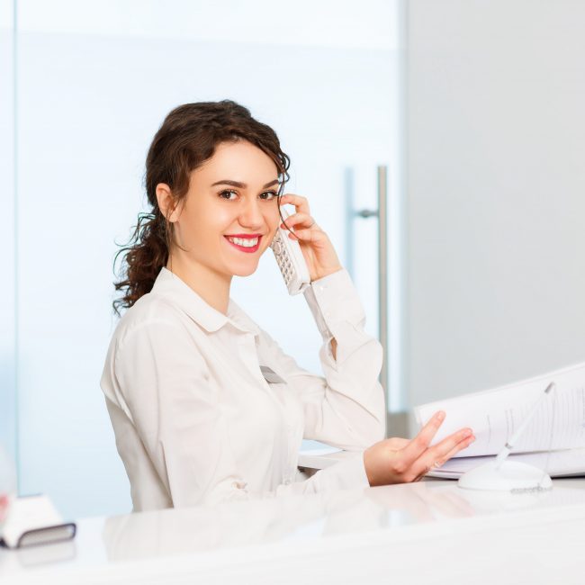 Friendly,Young,Woman,Behind,The,Reception,Desk,Administrator